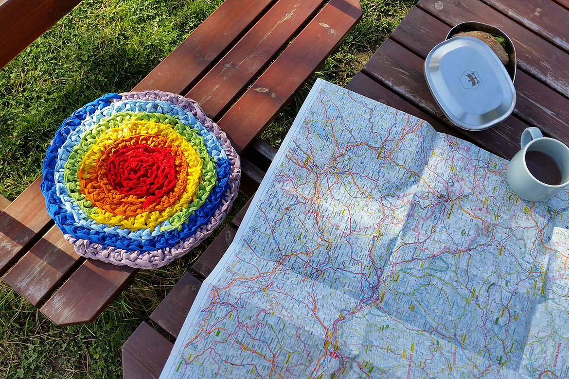 Das leuchtend bunte gehäkelte Upcycling-Sitzkissen liegt auf der Bank, auf dem Tisch liegen Landkarte und Vesper.