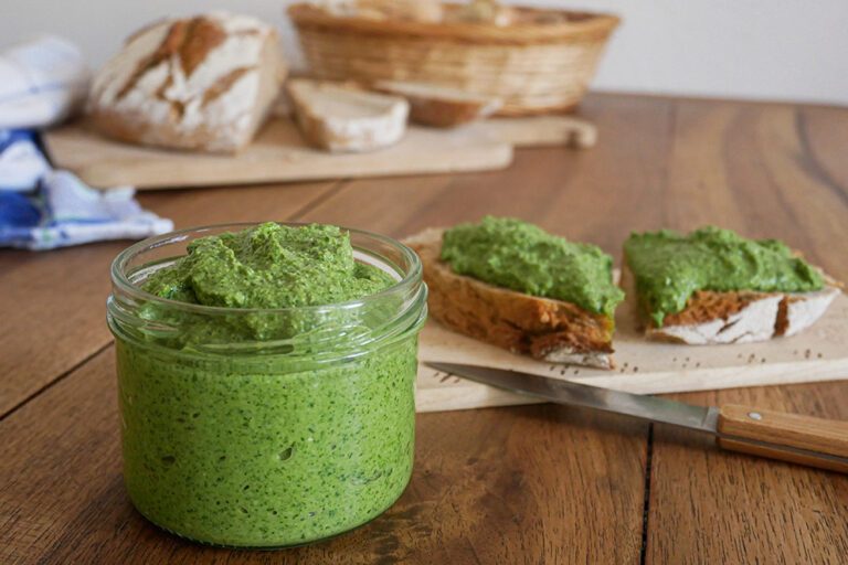 Das Koriander-Pesto steht in einem Glas auf einem Holztisch, auf dem zwei mit Pesto bestrichenen Brotscheiben liegen.