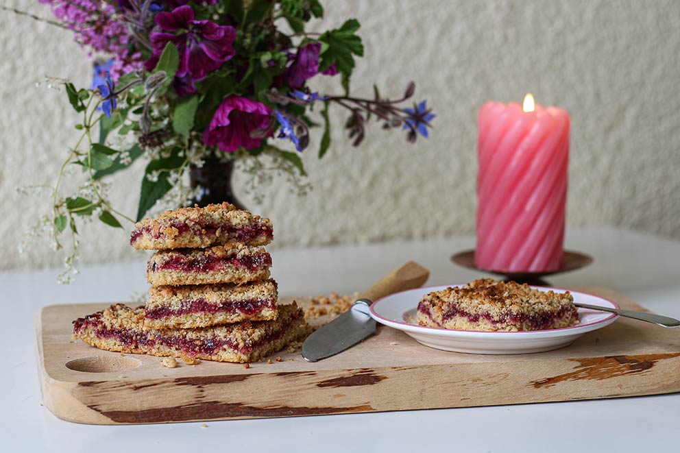 Der Marmeladenkuchen liegt in Stücke geschnitten auf einem Holzbrett, neben dem eine Kerze und Blumen in einer Vase stehen.